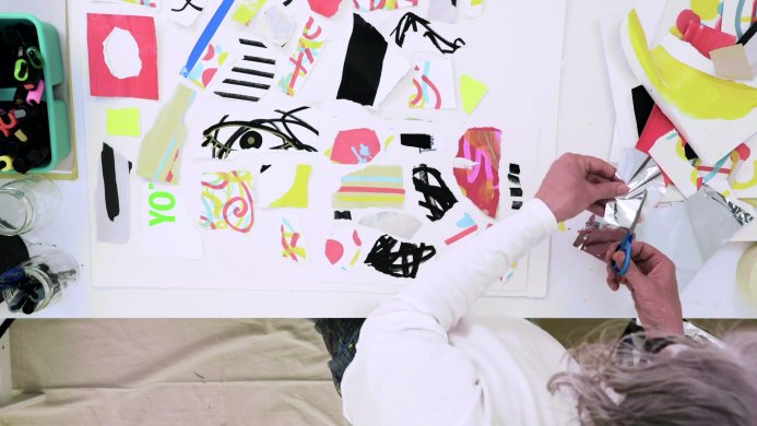 A bird's eye view of a man with gray hair cutting a piece of silver mylar to place on an artwork on the table in front of him