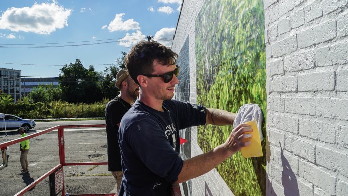A man rubbing a yellow sponge over a mural of trees affixed to a brick wall painted white